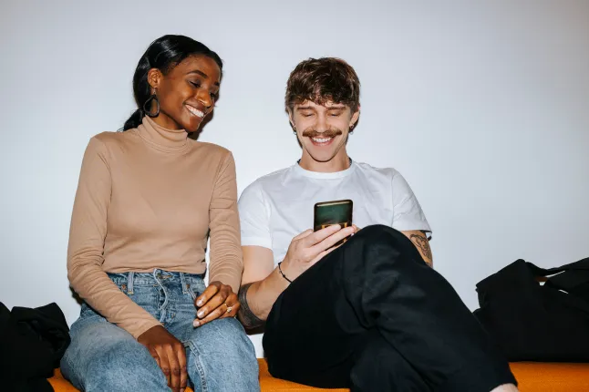 Õnnelik noormees, kes kasutab kolledžis valge seina lähedal koos meessõbraga diivanil istudes nutitelefoni