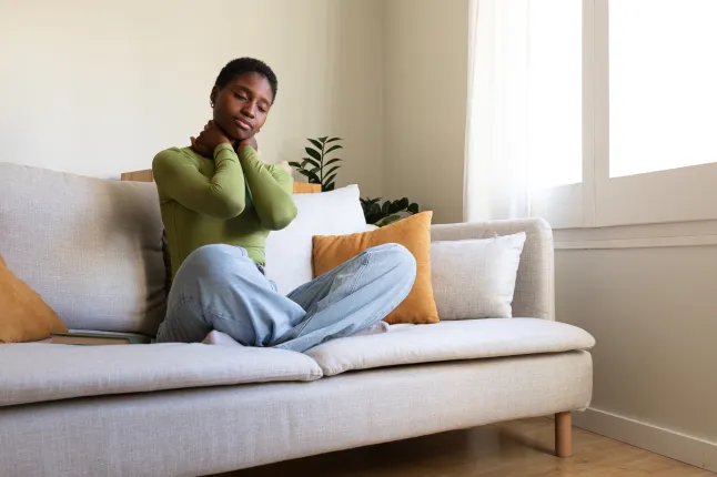 Noor afroameeriklanna istub diivanil, masseerides kaela. Mustanahaline naine kannatab kaelavalu all.