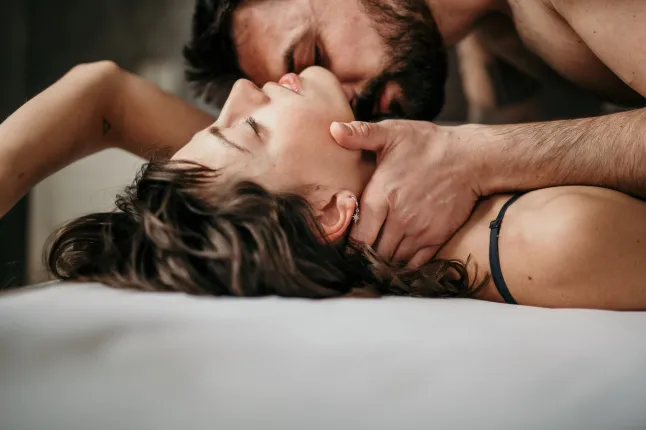 Külgvaate foto ilusast paarist voodis, olles õrn ja sensuaalne. Ilus habemega kutt kauni brüneti peal suudleb tema kaela, kui ta silmad on suletud. Armastuse kontseptsioon